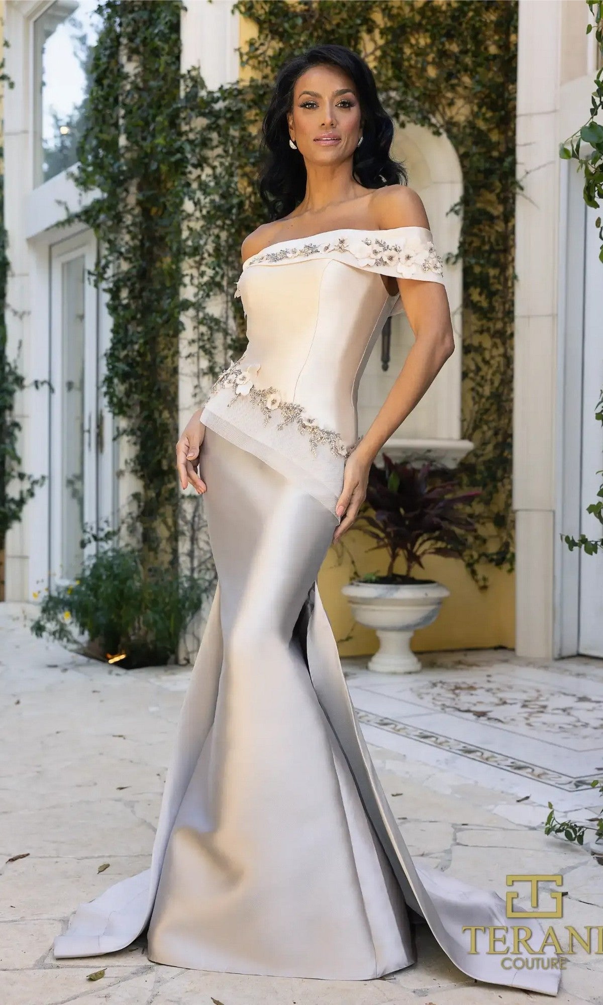 A woman in a champagne taupe Terani 242M3356 long formal dress stands outdoors near a vine-covered wall. The elegant gown is off-the-shoulder, mermaid Mikado style with floral embellishments.